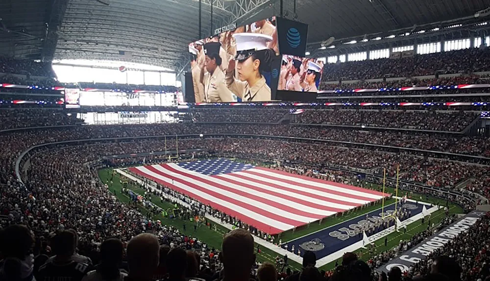 The image shows a packed stadium with a large American flag spread across the field while people in military uniforms are displayed on the big screens suggesting a patriotic event or sports game opening ceremony