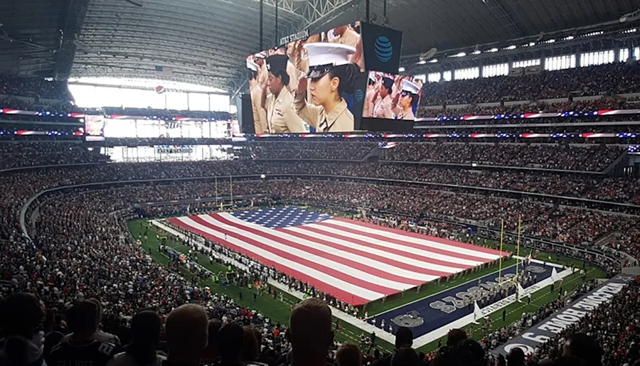 The image shows a packed stadium with a large American flag spread across the field, while people in military uniforms are displayed on the big screens, suggesting a patriotic event or sports game opening ceremony.