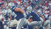 Players in football uniforms are poised for action during a game, with a focus on the offensive line and quarterback preparing for the next play.