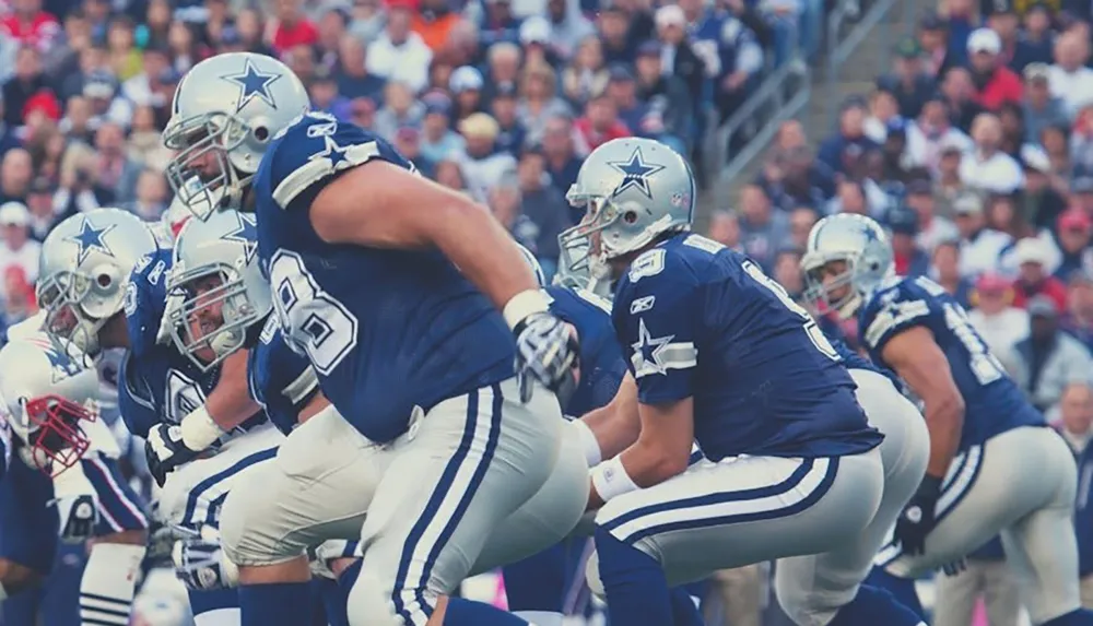 Players in football uniforms are poised for action during a game with a focus on the offensive line and quarterback preparing for the next play