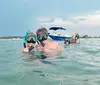 Two people wearing snorkeling gear give a thumbs-up in clear shallow water with boats and a beach in the background