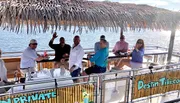 A group of people is enjoying a boat ride on a sunny day with a straw-covered overhang, waving and smiling at the camera.