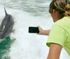 A person in a lime green shirt is taking a photo of a dolphin surfing a wave