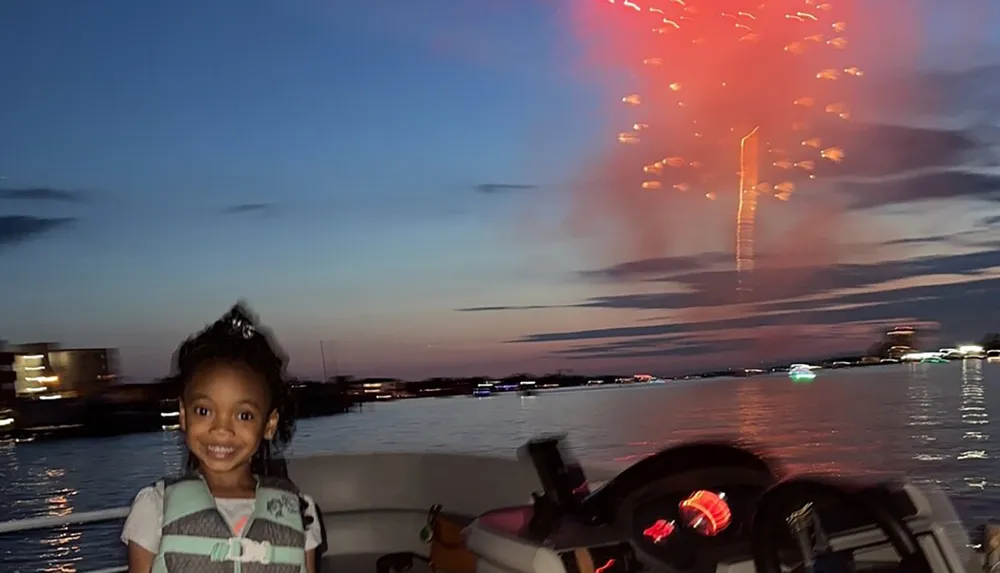 A young child is smiling on a boat with a backdrop of a colorful evening sky lit by fireworks