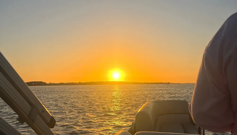 The image captures a serene sunset view over a body of water from the perspective of a boat with a part of a persons back in the foreground on the right