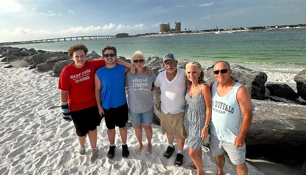 A group of six people possibly a family are smiling for a photo on a sandy beach with a jetty and buildings in the background