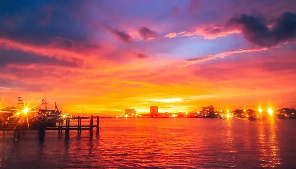 The image captures a vibrant and colorful sunset over a waterfront with silhouettes of boats and a pier against a backdrop of illuminated buildings