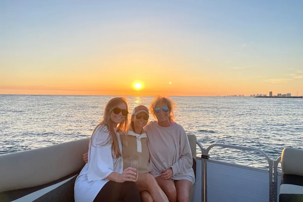 Three people are smiling and posing for a photo on a boat with a beautiful sunset over the water in the background