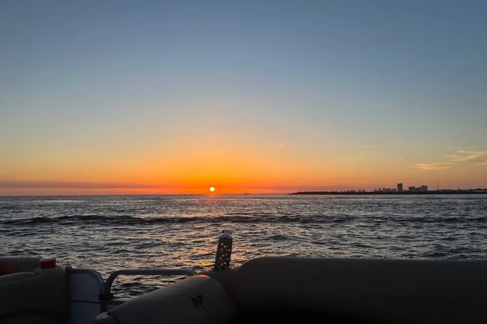 The photo captures a serene sunset over the ocean viewed from the side of a boat with a coastline visible in the distance