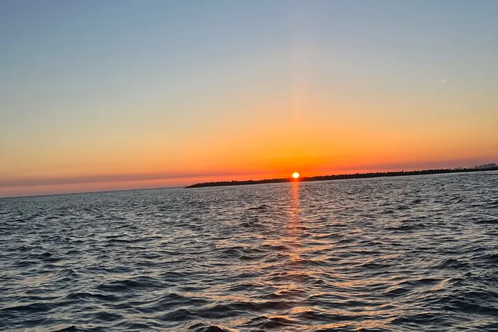 The image captures a tranquil sunset with the sun dipping near the horizon over a wavy sea painting the sky in hues of orange and blue