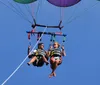 Two people are wearing life jackets and safety harnesses smiling and waving while enjoying a parasailing adventure over the water