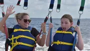 Two people are wearing life jackets and safety harnesses, smiling and waving while enjoying a parasailing adventure over the water.
