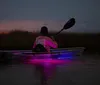 A person is kayaking in tranquil waters at dusk with a striking purple light illuminating the water beneath the kayak