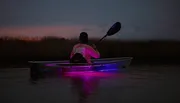 A person is kayaking in tranquil waters at dusk with a striking purple light illuminating the water beneath the kayak.