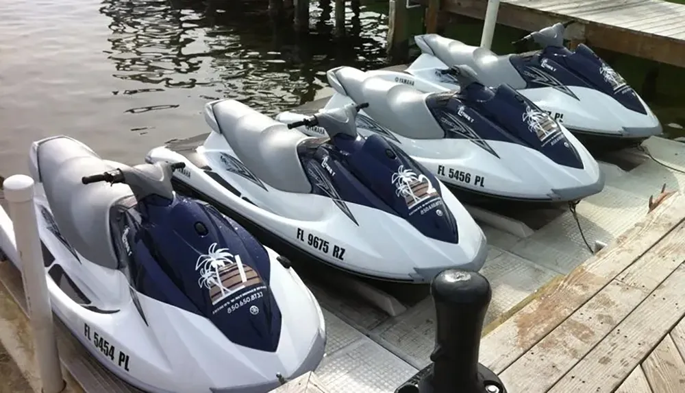 The image shows a row of four identical white and blue personal watercrafts docked at a wooden pier on a body of water