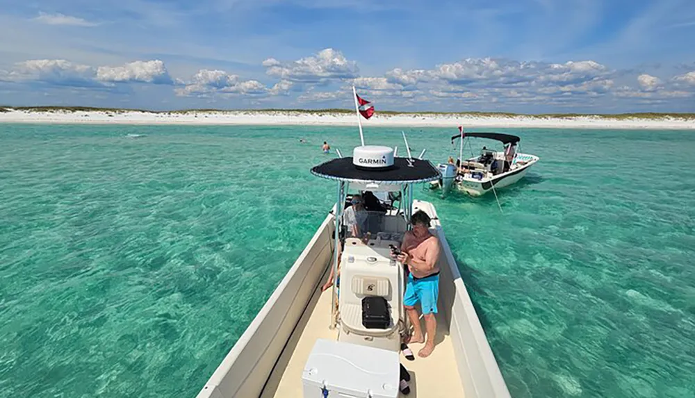 A person stands on a boat floating in clear turquoise waters near a sandy beach with other boats and people enjoying the day