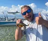 A man gives a thumbs-up while happily displaying a small fish in front of a boat anchored in shallow waters