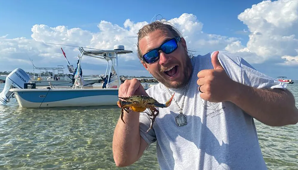 A man gives a thumbs-up while happily displaying a small fish in front of a boat anchored in shallow waters