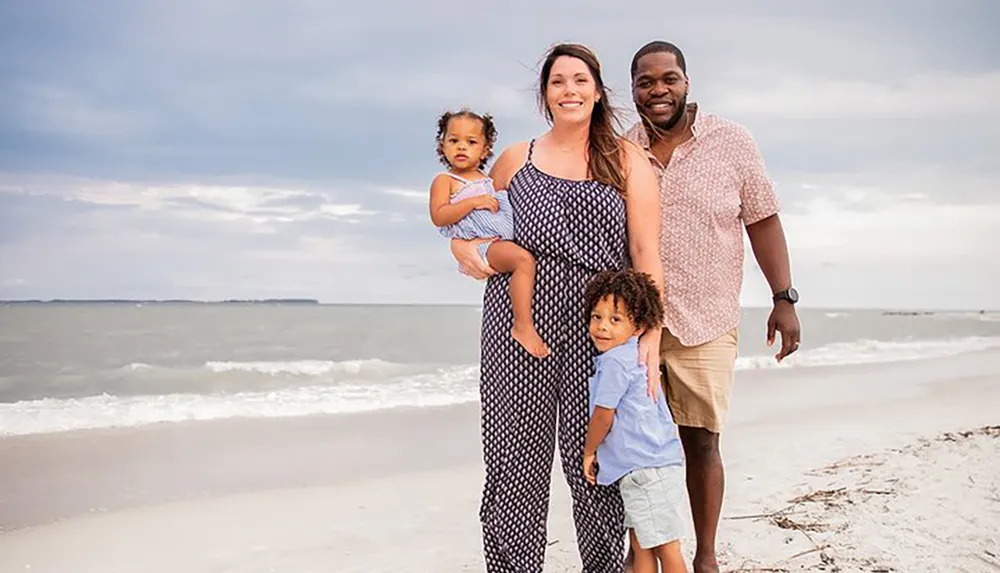 A happy family with two young children is standing on a beach smiling and posing for a photo with the ocean in the background