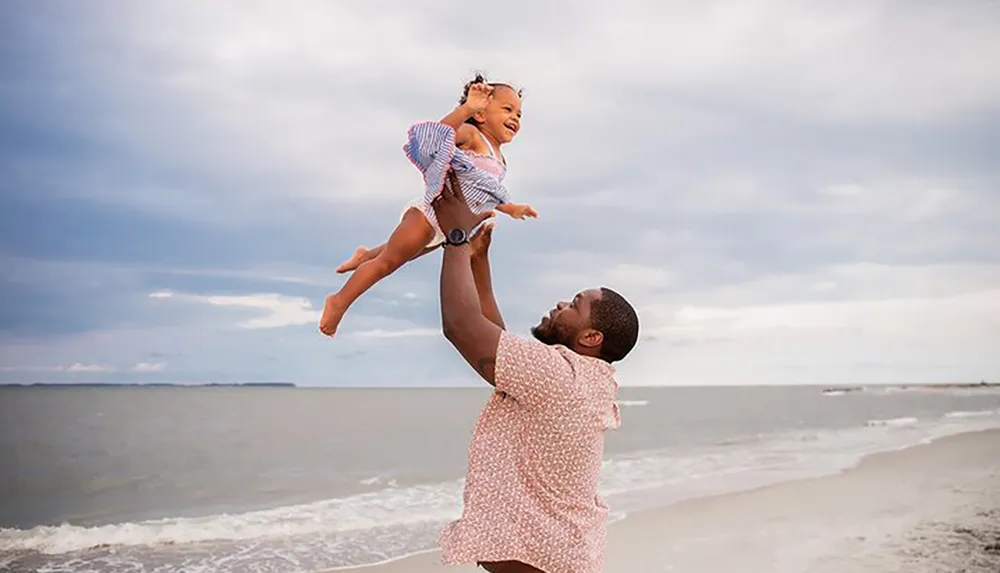 A joyful child is being lifted into the air by an adult on a beach under a cloudy sky