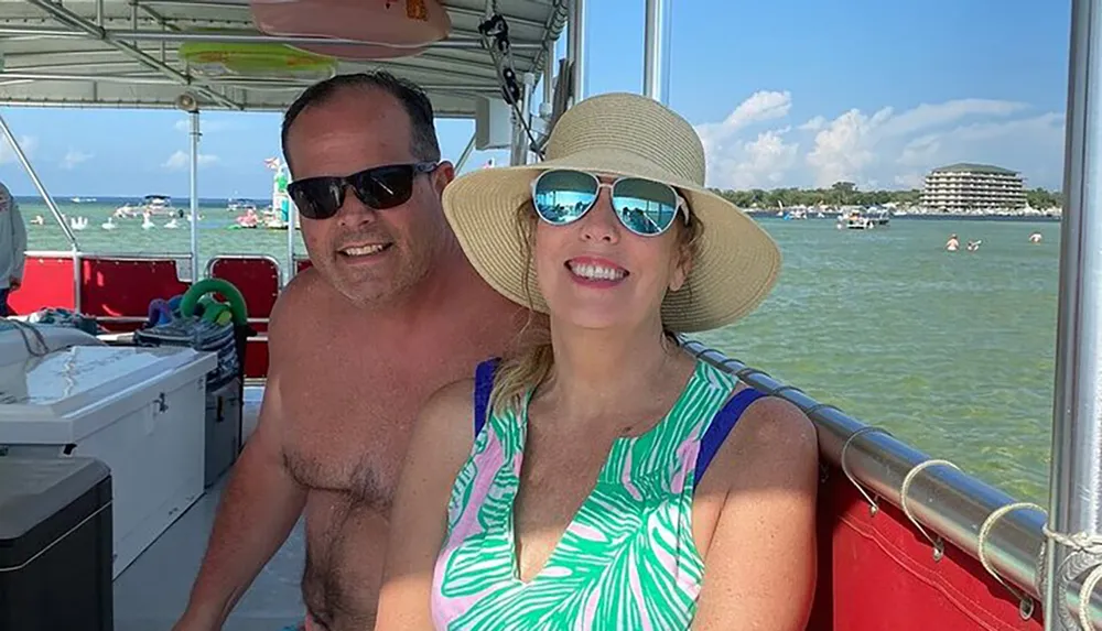 A man and a woman are smiling for a selfie on a sunny day aboard a boat with a lake and other boaters in the background