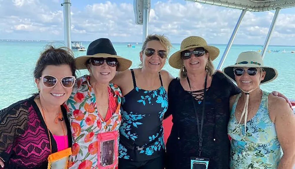 Five smiling women are posing together against a backdrop of clear blue water on a sunny day all wearing sunglasses and some wearing hats suggesting a leisurely gathering or vacation