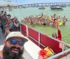 A group of people is joyfully posing for a selfie on a boat while another crowd in the water behind them is also partaking in the photo all set against a backdrop of a sunny busy waterway filled with boats