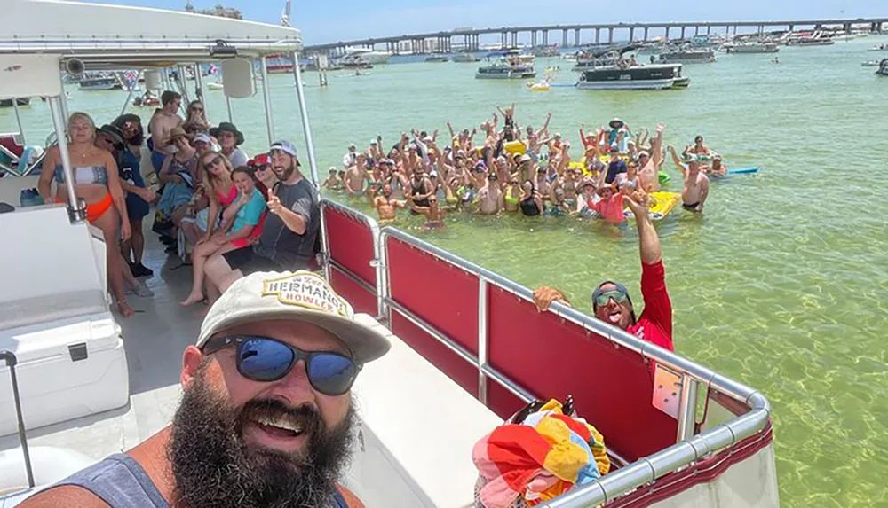 A group of people is joyfully posing for a selfie on a boat while another crowd in the water behind them is also partaking in the photo all set against a backdrop of a sunny busy waterway filled with boats