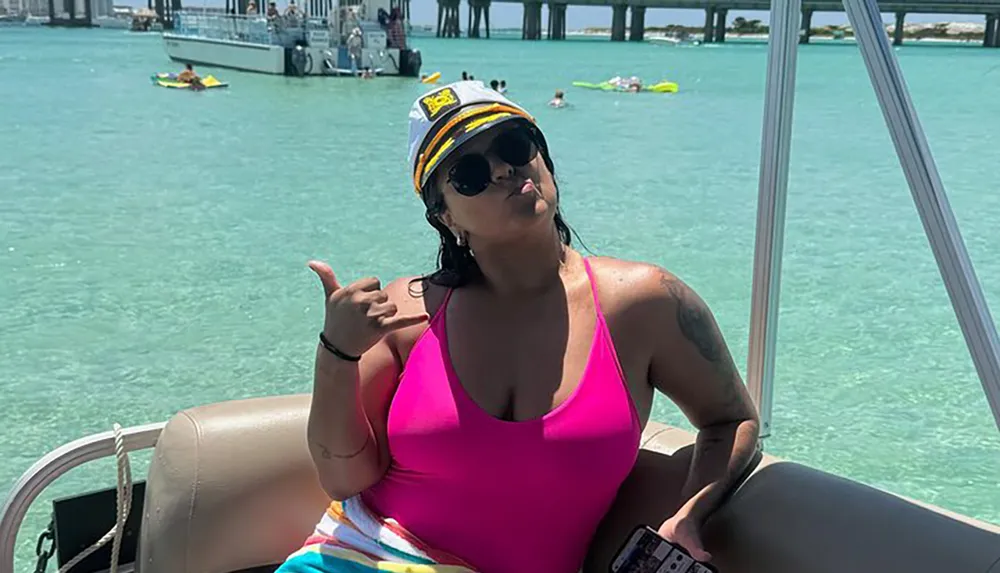 A person is posing with a shaka hand gesture wearing a captains hat and sunglasses on a boat with aquamarine waters and other watercraft in the background