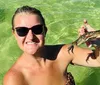 A smiling woman in sunglasses is standing in clear green water holding a small crab up to the camera