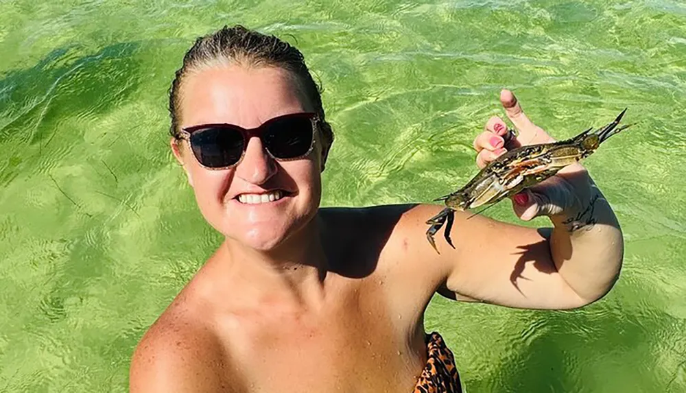 A smiling woman in sunglasses is standing in clear green water holding a small crab up to the camera