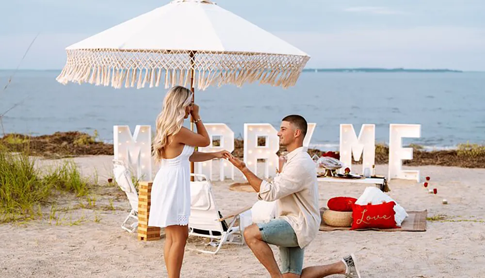 A person is proposing on a sandy beach with a romantic Marry Me sign and decorations in the background