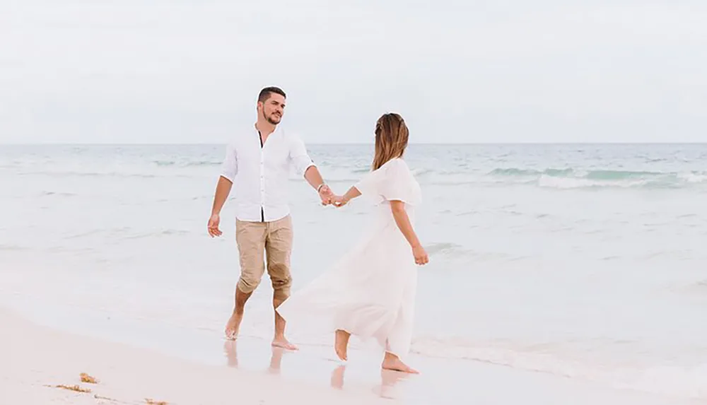 A couple holds hands while walking on a sandy beach with the ocean in the background