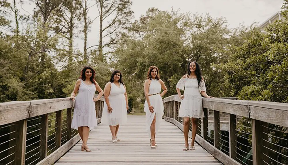 Four women dressed in white outfits are standing spaced apart on a wooden boardwalk amidst a lush green environment