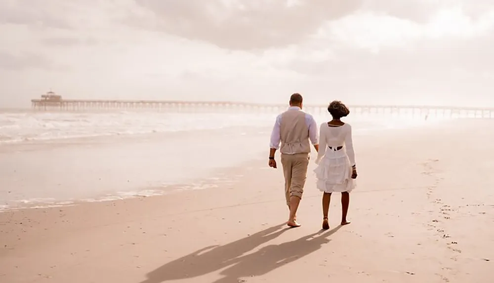 A couple is walking together hand in hand along a sunlit beach facing away from the camera with a pier in the background