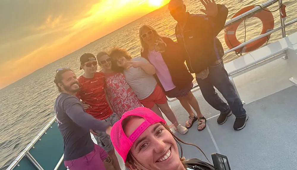 A group of people is posing for a selfie on a boat enjoying a sunset over the sea