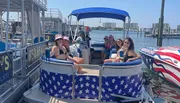 A group of smiling people is enjoying a sunny day on a boat with patriotic American flag-themed covers on the seats.