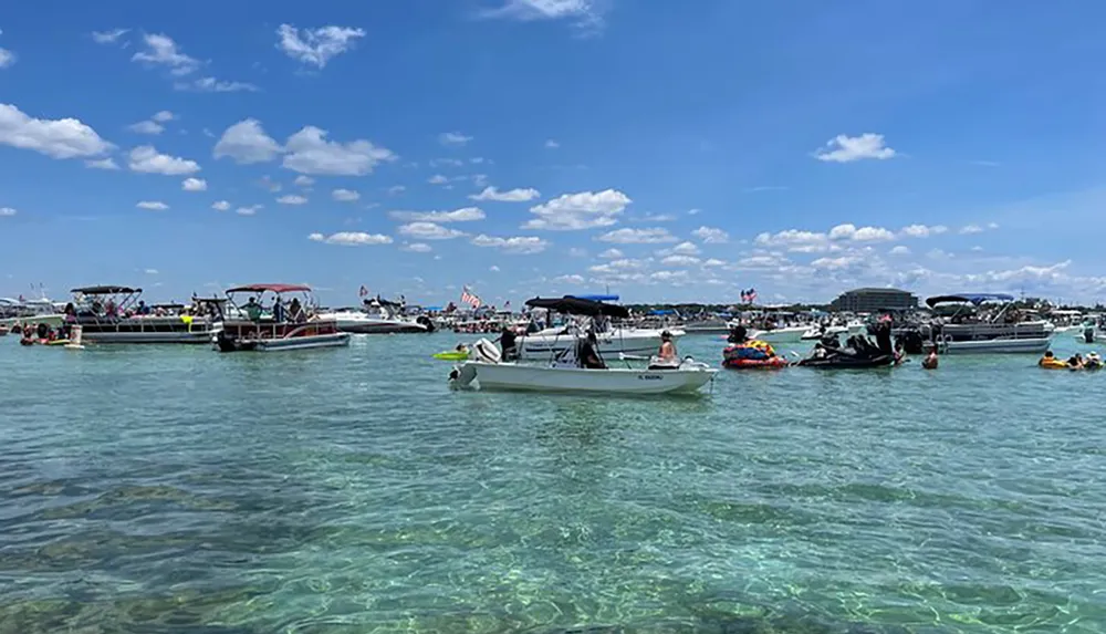 A lively gathering of people enjoying a sunny day on the water with various boats and inflatables anchored in a clear turquoise bay