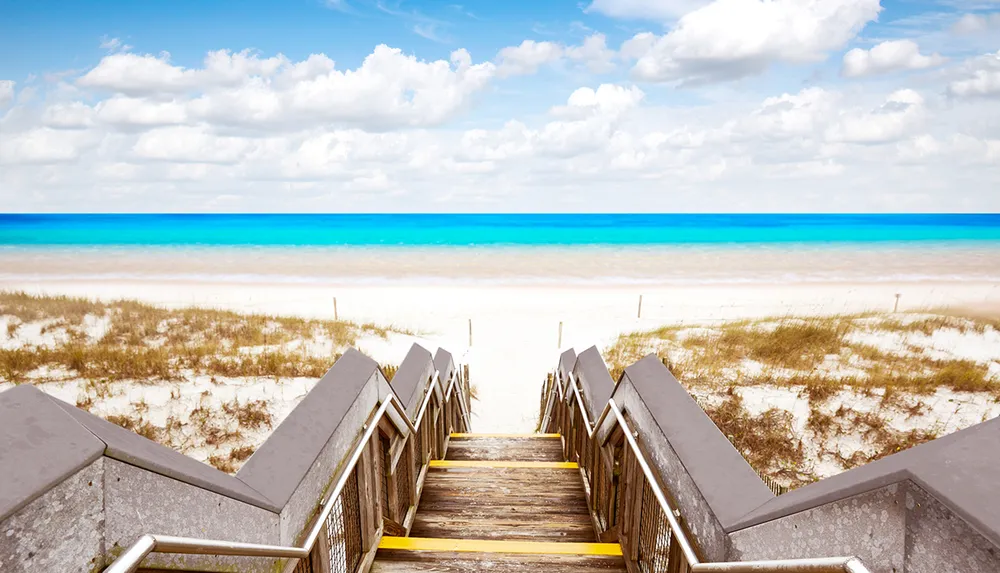 A wooden stairway leads down to a serene white sandy beach with a clear turquoise ocean and blue sky dotted with white clouds