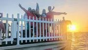 A group of people is posing happily behind a white picket fence on a waterfront at sunset.