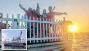 A group of people is posing happily behind a white picket fence on a waterfront at sunset.