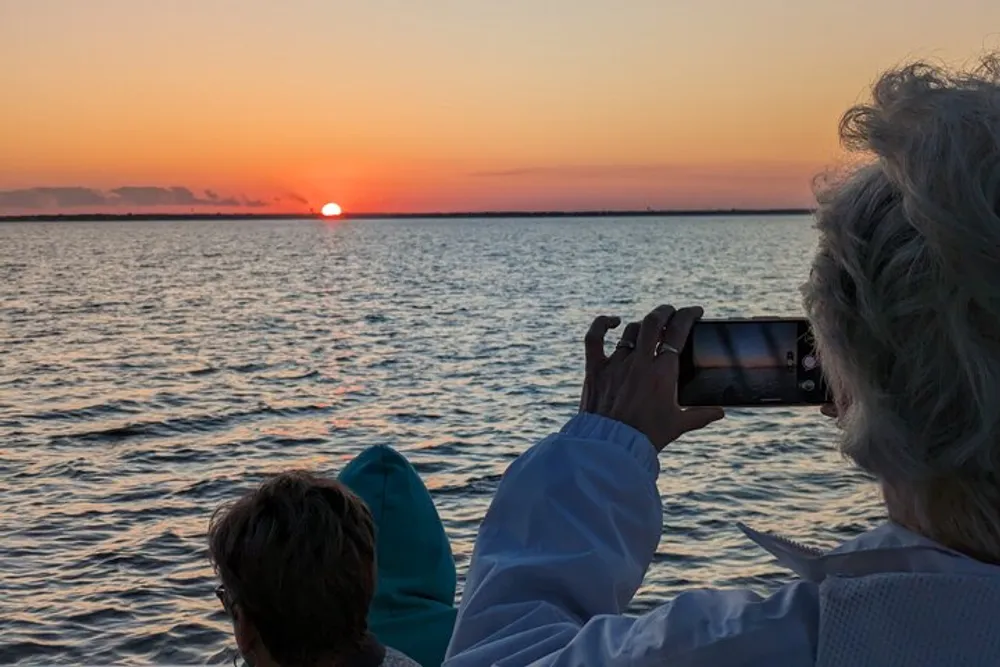 A person is capturing the sunset over the sea with their smartphone while another person sits beside them