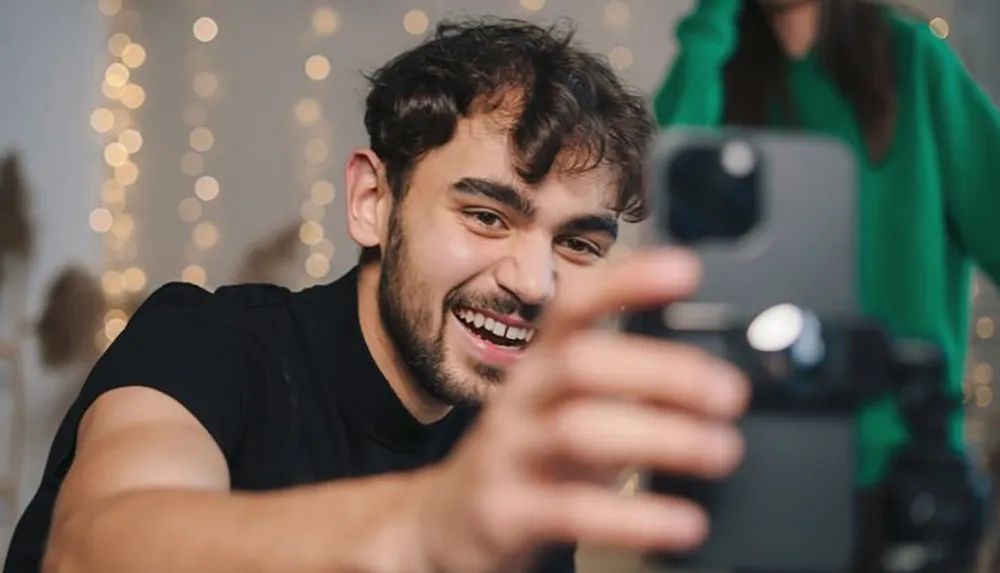 A man with a cheerful expression is taking a selfie with a smartphone attached to a tripod with a blurred person in the background and bokeh lights enhancing the festive atmosphere