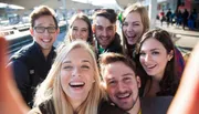 A group of cheerful young adults is taking a selfie with one of them holding the camera out in front, capturing their smiling faces.