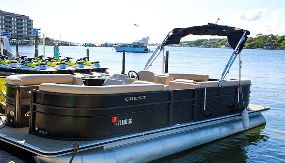 A pontoon boat is docked by the waterfront with jet skis and other boating activities visible in the background