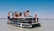 A group of young people are enjoying a sunny day on a pontoon boat, waving and smiling, with clear blue skies and water in the background.