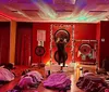 The image shows a dimly-lit room with a cosmic star projector effect on the ceiling while several people lay on mats under blankets and a person at the front appears to be performing a sound healing or meditation session with large gongs