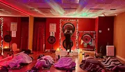 The image shows a dimly-lit room with a cosmic star projector effect on the ceiling, while several people lay on mats under blankets, and a person at the front appears to be performing a sound healing or meditation session with large gongs.