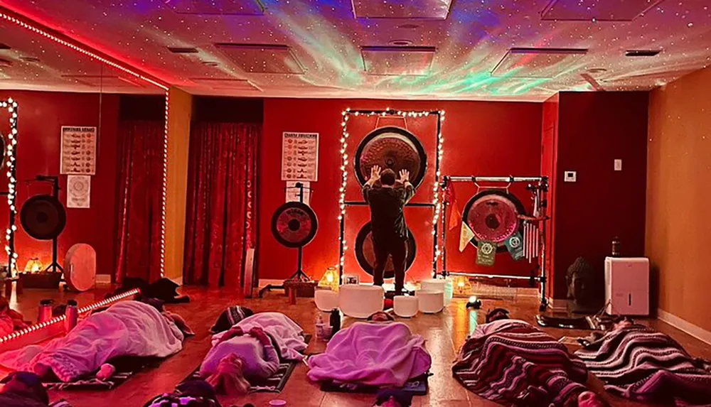 The image shows a dimly-lit room with a cosmic star projector effect on the ceiling while several people lay on mats under blankets and a person at the front appears to be performing a sound healing or meditation session with large gongs