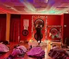 The image shows a dimly-lit room with a cosmic star projector effect on the ceiling while several people lay on mats under blankets and a person at the front appears to be performing a sound healing or meditation session with large gongs